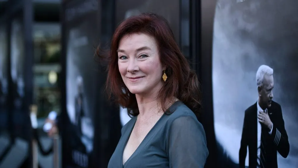 Woman with red hair wearing a gray dress and earrings, smiling at an event.