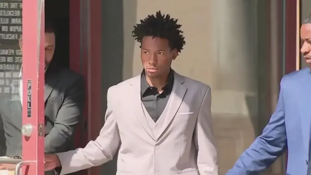 Young man wearing a gray suit and black shirt exiting a building with two other individuals.