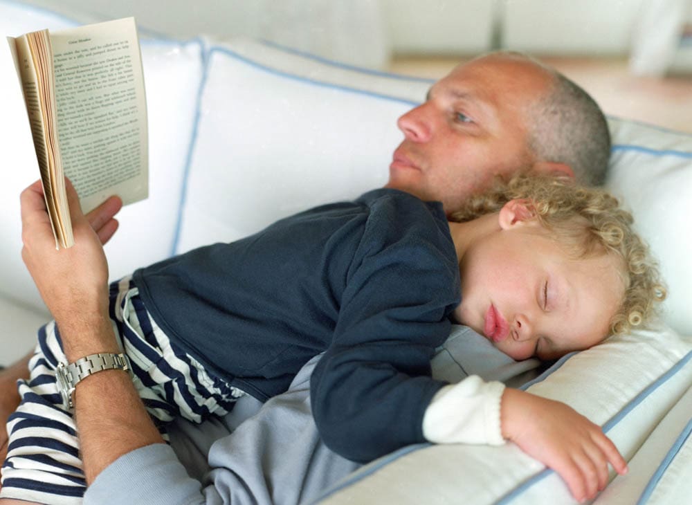 A man reading a book while a sleeping child rests on his chest in a cozy setting.