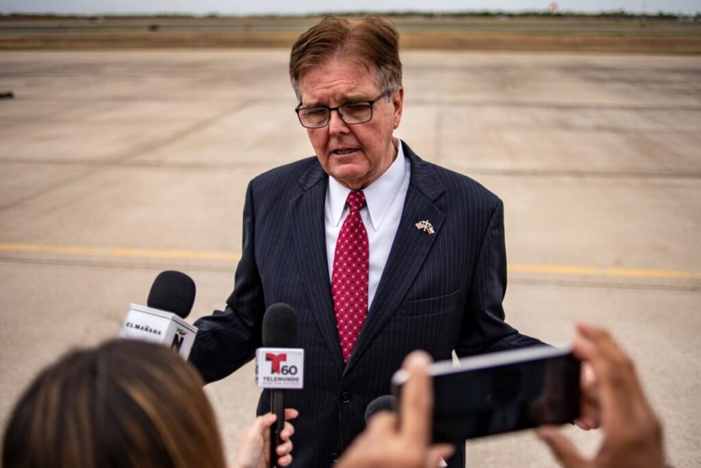 A man in a suit with a red tie speaking to reporters at an airport, surrounded by microphones.