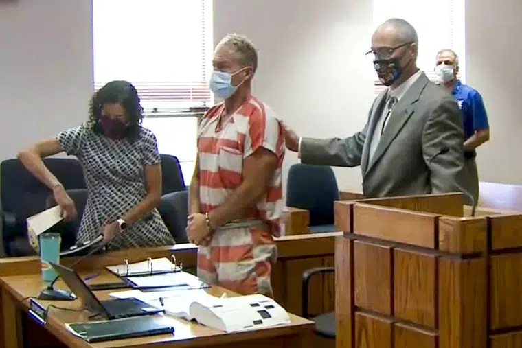 Man wearing an orange jail uniform standing in a courtroom with two individuals nearby.