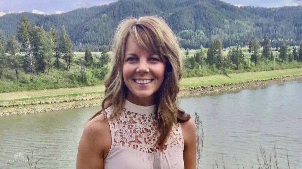 A woman with long brown hair smiling near a river surrounded by trees and mountains.