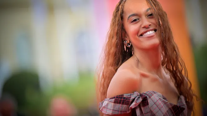 Cindy Bruna smiling while wearing a plaid off-shoulder dress at an event.
