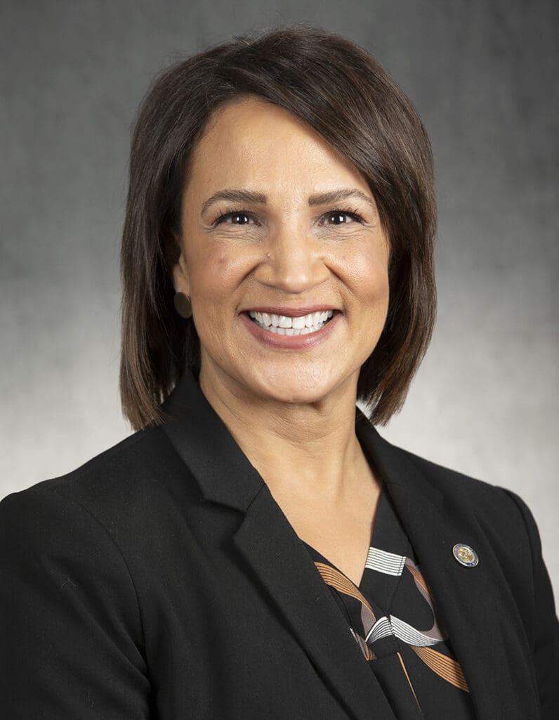 Professional headshot of a woman with shoulder-length brown hair wearing a black blazer and smiling at the camera.