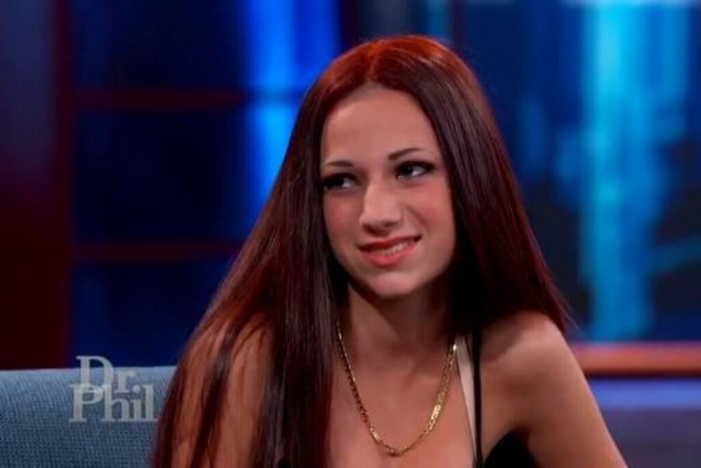Young woman with long brown hair and a chain necklace sitting on a television set.