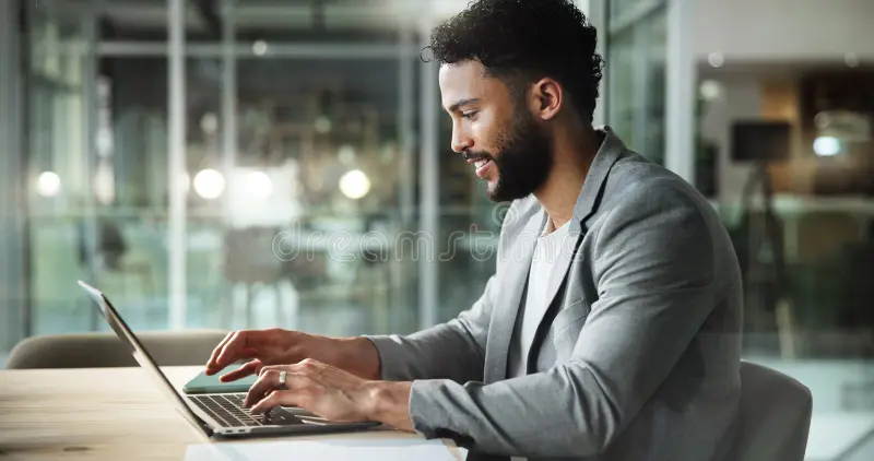 Man in a gray suit typing on a laptop in an indoor setting with soft lighting.