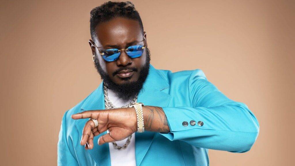 T-Pain wearing a blue suit and sunglasses, checking his watch with a serious expression.
