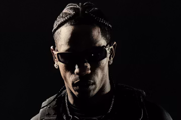 Portrait of Travis Scott wearing sunglasses and a dark outfit against a black background.