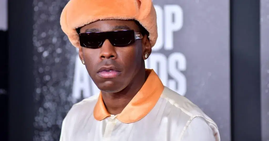 Tyler, the Creator wearing sunglasses and an orange hat at a formal event.