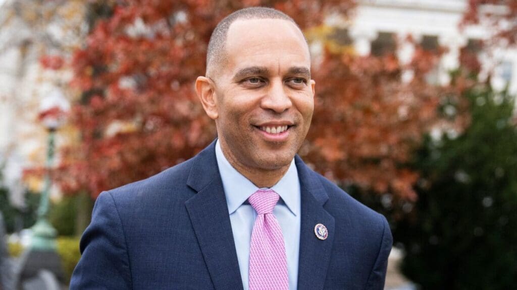 Hakeem Jeffries wearing a suit and pink tie, smiling outdoors with trees in the background.