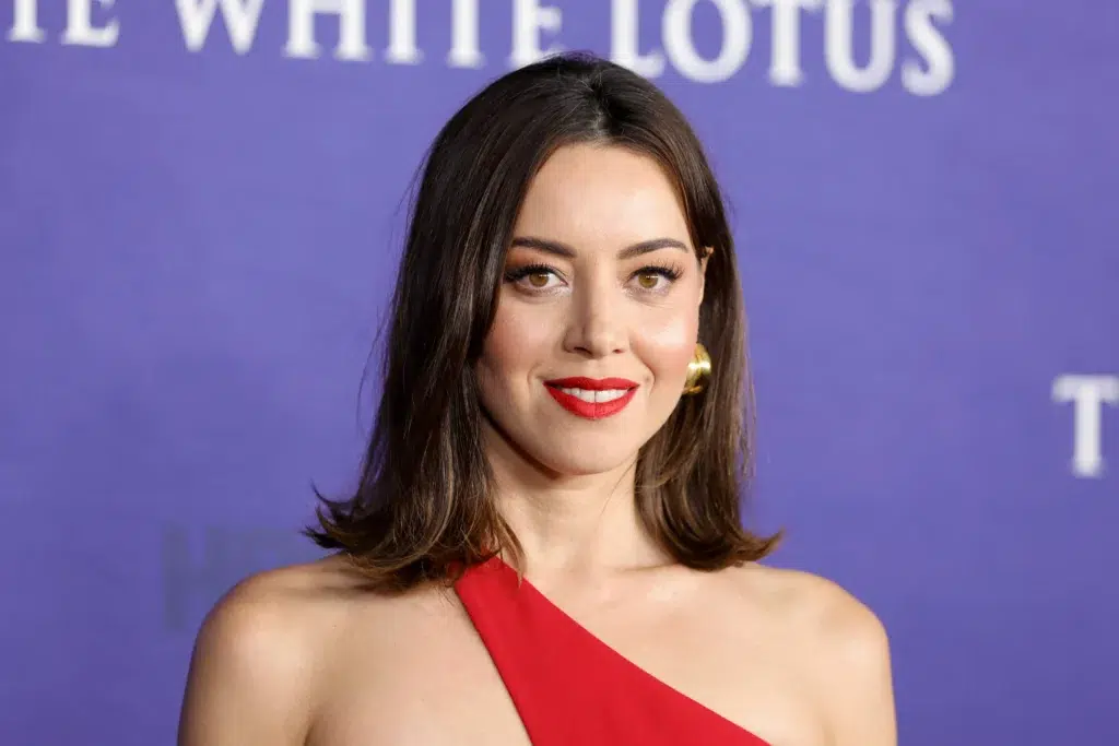 Aubrey Plaza posing for a photo at The White Lotus event with a red dress and bold lipstick.